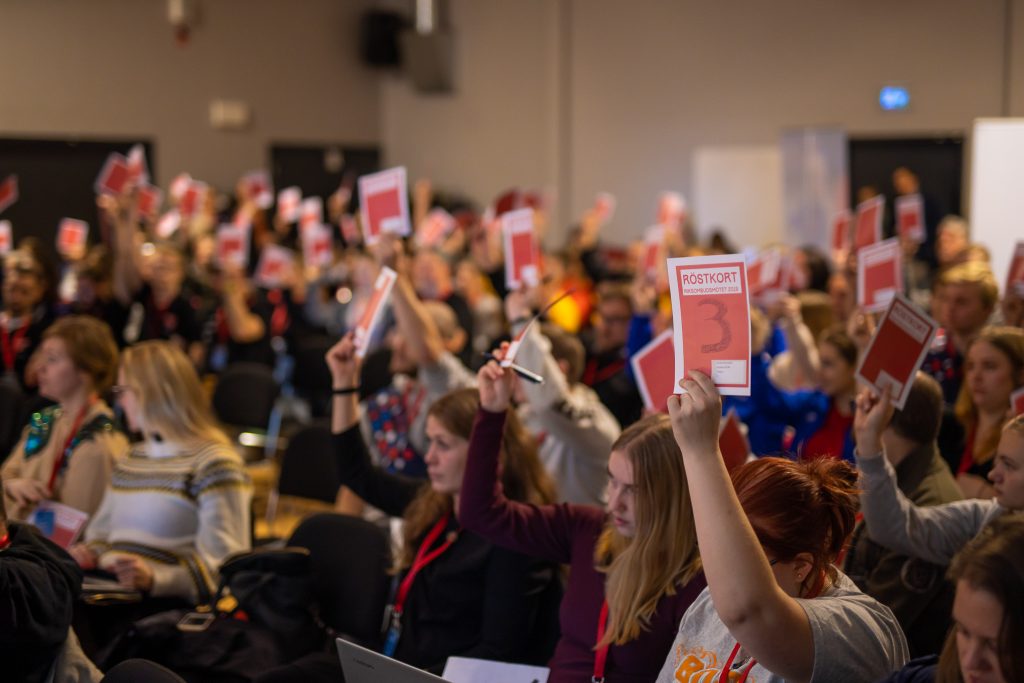 Deltagare på KFUMs riksombudsmöte 2023 röstar genom att höja sina röstkort, exemplifierar årsmötesprocessen inom KFUM, bifogad som referens på sidan för KFUM Ängsholmens årsmöte 2023.